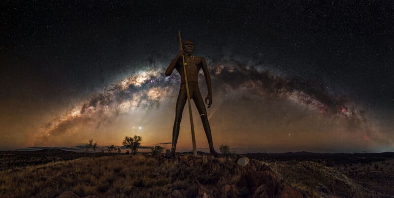 A tall statue of an Aboriginal man holding a spear stands on a hill under a night sky, with the Milky Way galaxy arching brightly overhead. Sparse vegetation and distant hills are visible on the ground.