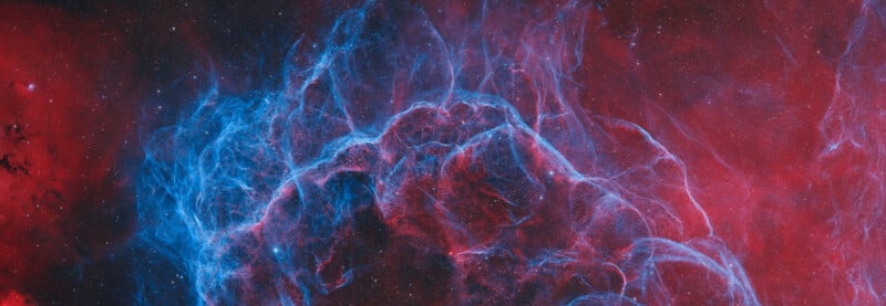 Vivid image of a nebula in deep space, featuring bright blue and red glowing gas clouds against a dark background scattered with tiny stars. The swirling colors create a dramatic, ethereal effect.