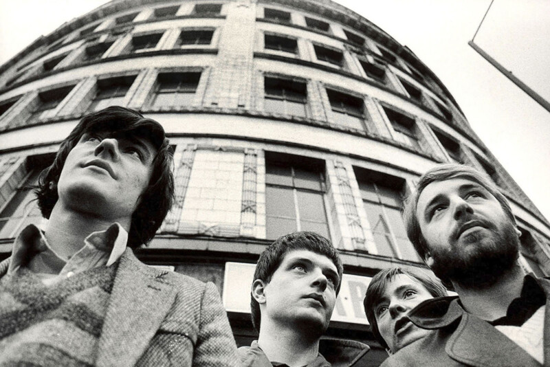 Four men stand close together, looking slightly upward, in front of a tall, curved building. The photo is black and white, taken from a low angle, emphasizing the building’s facade and the serious expressions on their faces.