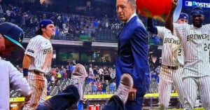 A TV broadcast shows a baseball field with players in Brewers uniforms celebrating. One man in a suit looks back while another person’s legs and shoes are visible in the foreground, as if being tossed or falling.