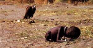 A malnourished child is crouched on dry, barren ground while a vulture stands nearby, watching. The scene highlights severe famine and suffering in a harsh environment.