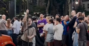 A crowd of people stands on a city street at dusk, many looking up and taking photos. A person in the foreground holds up a pole and seems excited, while others smile and observe the scene above.