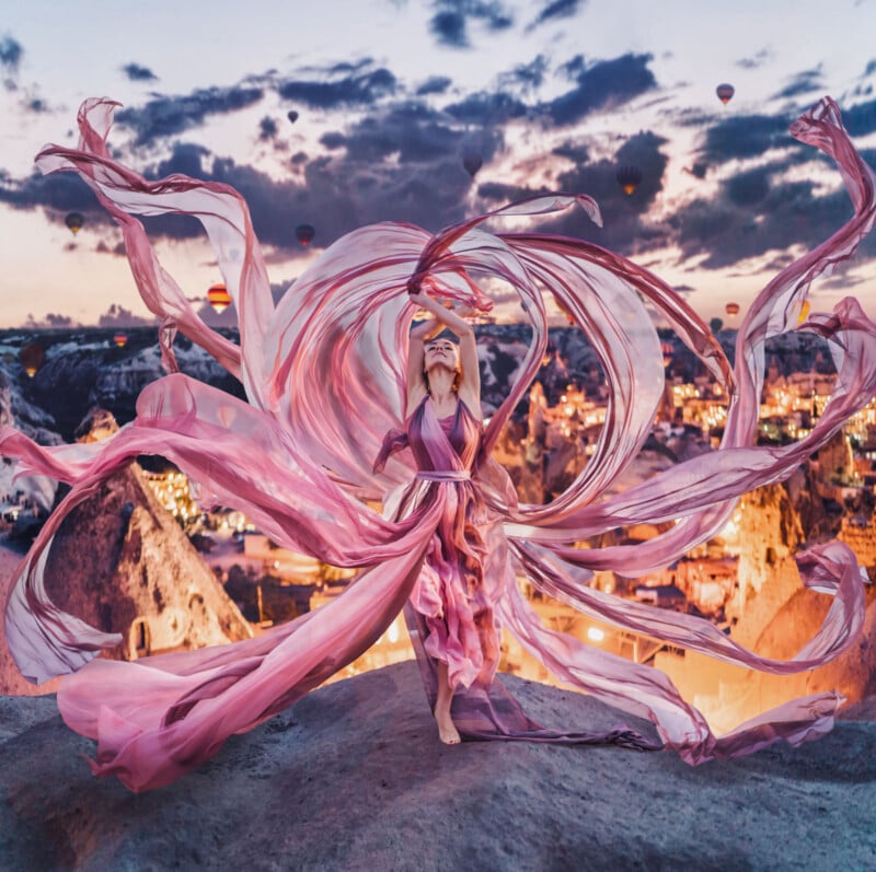 A woman in a flowing pink dress dances on a rock at sunset, with long fabric swirling around her and hot air balloons floating in the sky above a brightly lit town and rocky landscape.