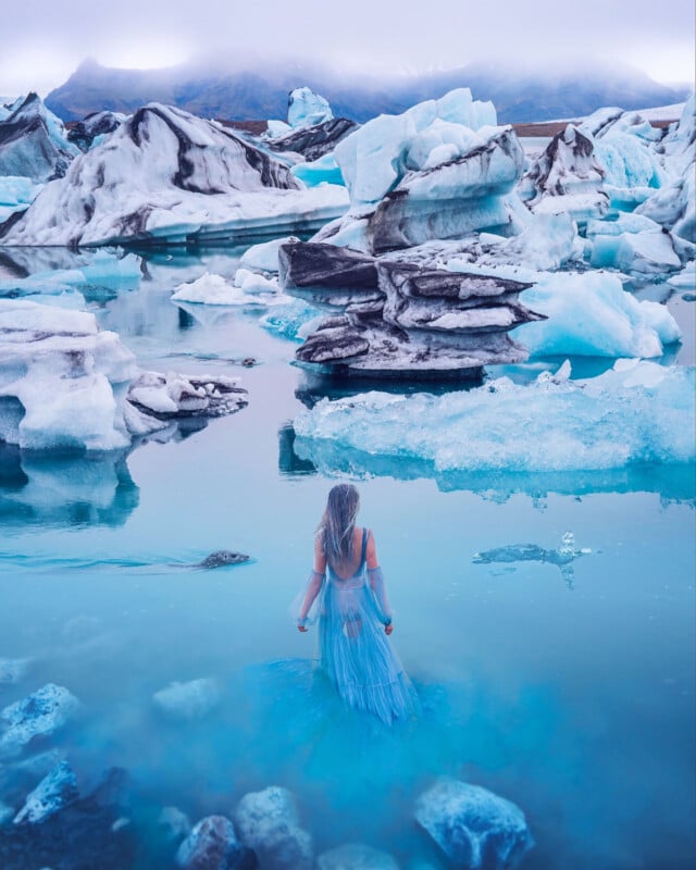 A woman in a long, flowing blue dress stands in icy water surrounded by large blue and white icebergs, with misty mountains in the background. The scene is serene and dreamlike.