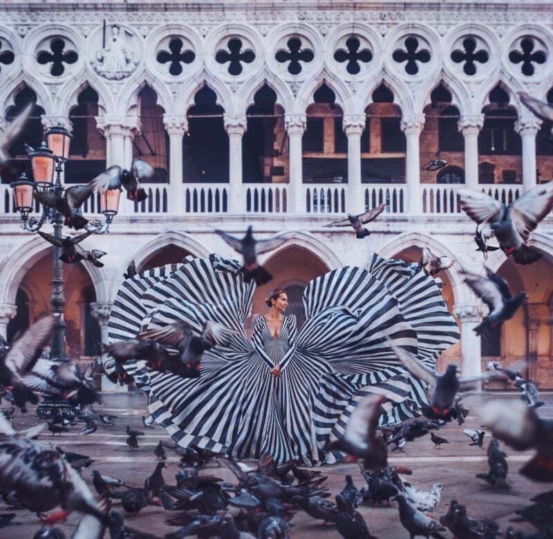 A woman in a dramatic, black-and-white striped dress stands in front of a historic building with arches, surrounded by flying pigeons. Her dress fans out in large, circular shapes, creating a striking visual effect.