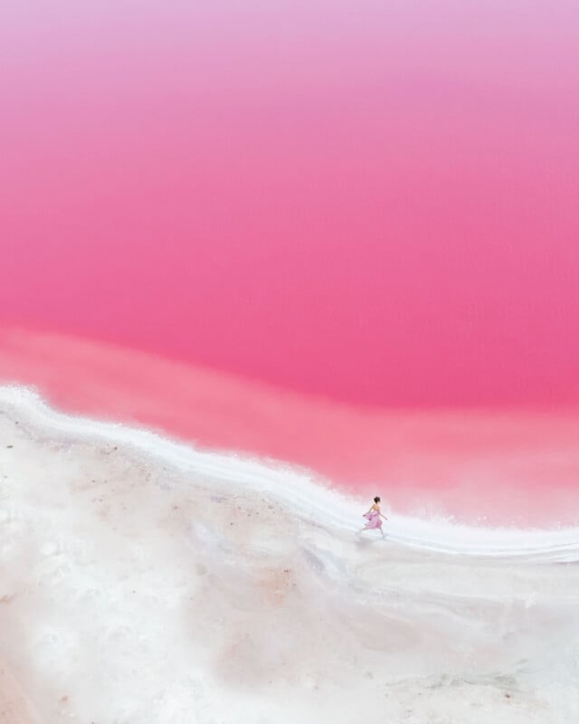A person in pink walks along the white sandy shore of a vibrant pink lake, captured from above, with the striking color contrast between the rosy water and pale shoreline.