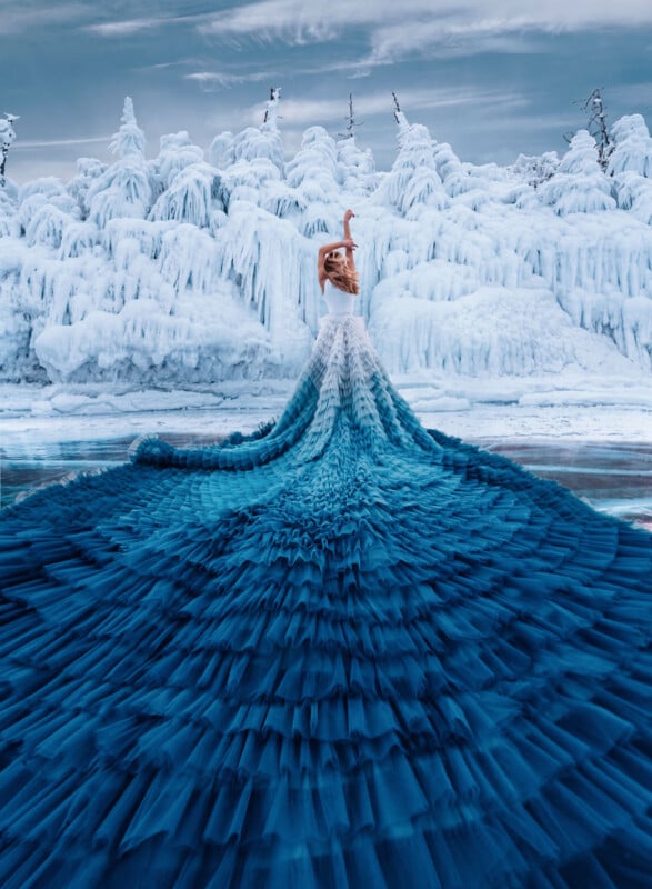 A woman in a long, flowing blue gown stands on icy ground before snow-covered trees and frozen landscape, her dress blending dramatically into the wintry scene under a cloudy sky.
