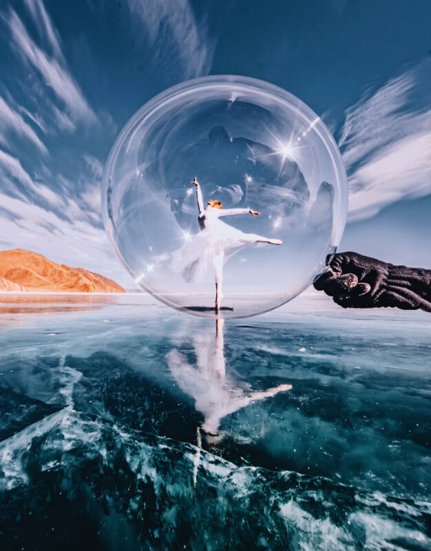 A ballerina in a white tutu poses gracefully inside a large transparent bubble on a frozen lake, with a gloved hand holding the bubble and dramatic mountains and sky in the background.