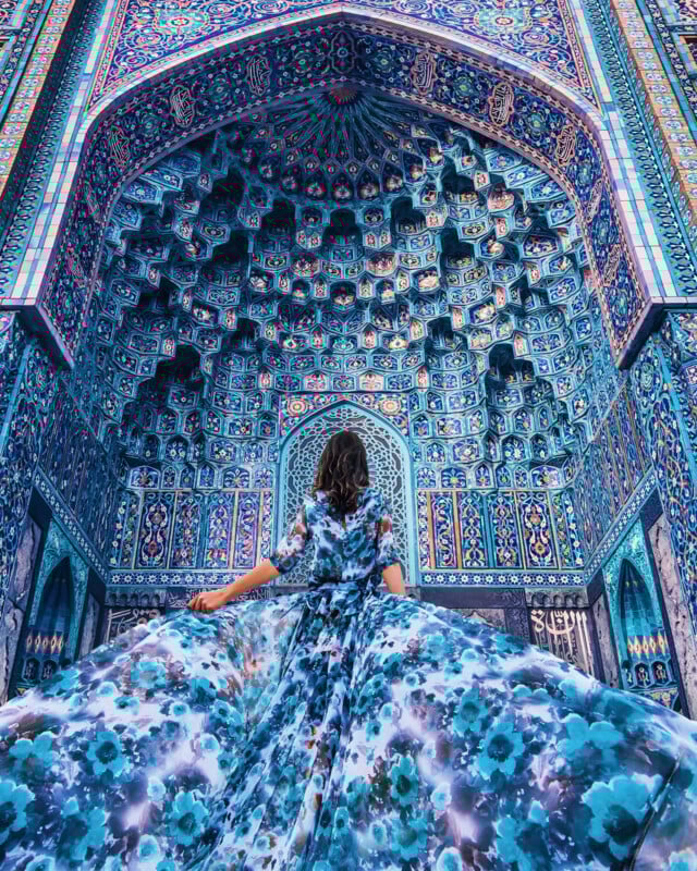 A woman in a flowing, blue floral dress stands facing an intricate, colorful mosaic archway, blending with the ornate blue and teal patterns of the Islamic architecture above her.
