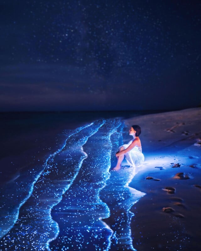 A woman in a white dress sits on a sandy beach at night, surrounded by glowing blue waves and twinkling lights, with starry skies above, creating a magical, surreal atmosphere.