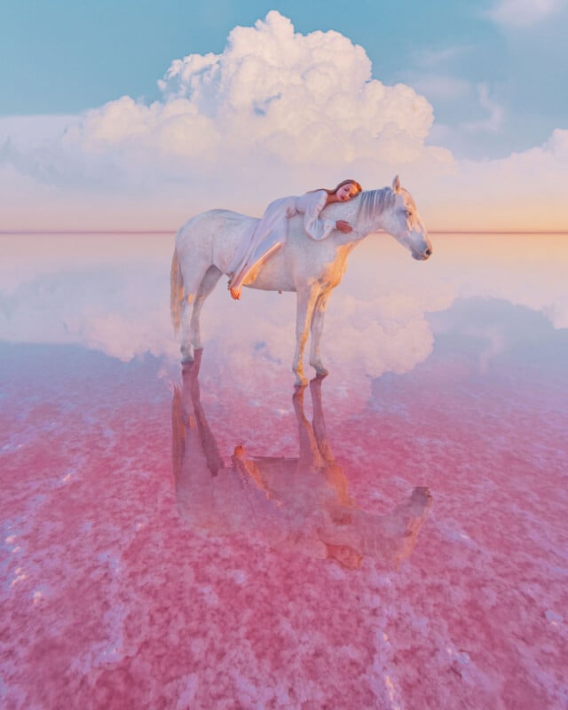 A woman in a pale dress lies across the back of a white horse, both reflected in pink, shallow water. Fluffy clouds and a pastel sky create a surreal, dreamy landscape.