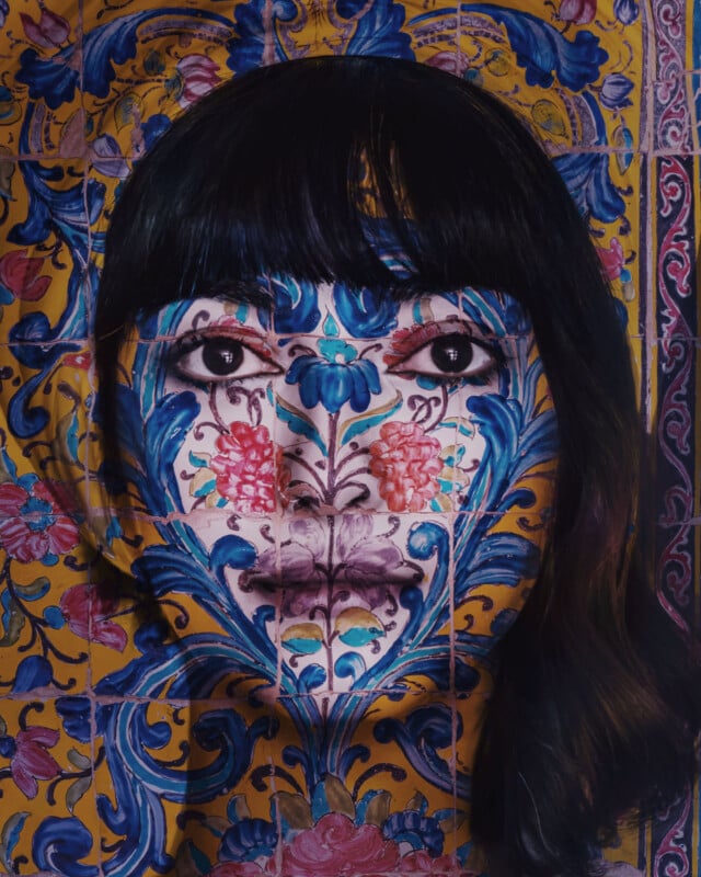 A woman's face is painted to blend seamlessly with a vibrant, intricate floral tile pattern in blue, pink, and yellow, creating an illusion where her features merge with the ornate background.
