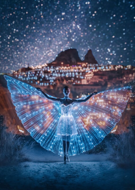 A woman stands outdoors at night wearing a glowing, butterfly-wing-shaped dress made of blue and white lights, with a starry sky and distant illuminated cityscape behind her.