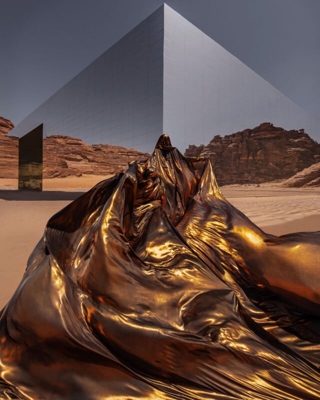 A large mirrored cube building reflects rocky desert surroundings. In the foreground, shiny metallic gold fabric is draped dramatically across the sand, creating a surreal, otherworldly scene.