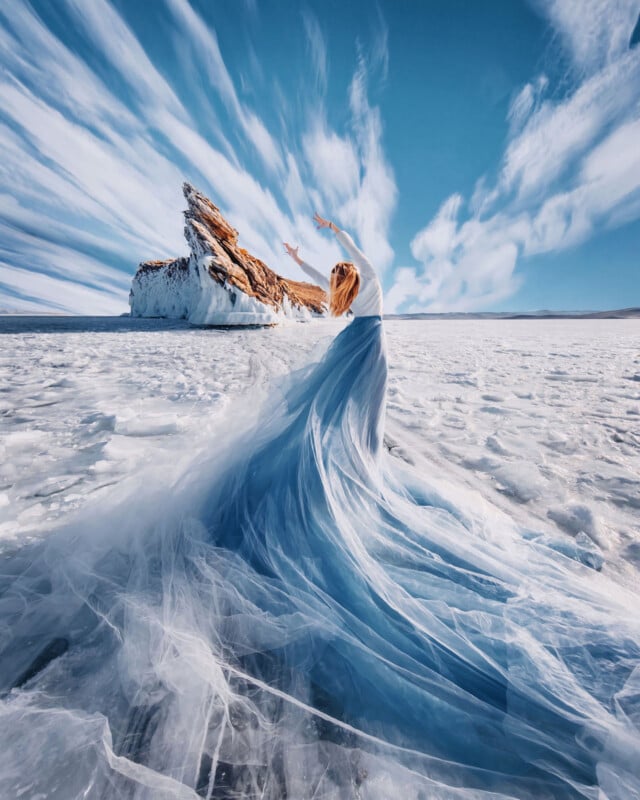 A woman in a flowing blue dress stands on a vast, frozen landscape, her arms raised toward the sky. Dramatic clouds streak above, and a rocky, ice-covered formation rises in the distance.