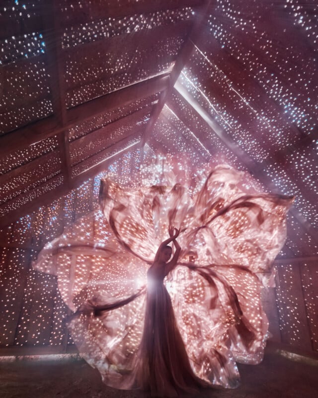 A person in a flowing dress poses gracefully inside a wooden structure with star-like holes, illuminated by pink and purple light, creating a magical, ethereal atmosphere with swirling fabric and sparkling light patterns.
