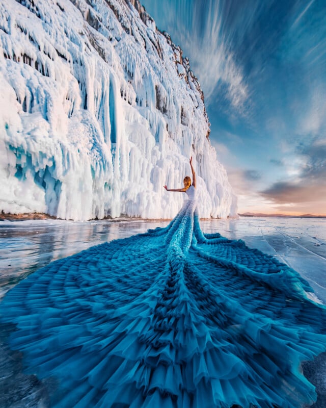 A person in a dramatic, flowing blue gown stands on icy ground near tall, frozen cliffs under a vibrant sky, creating a striking contrast with the snowy landscape.