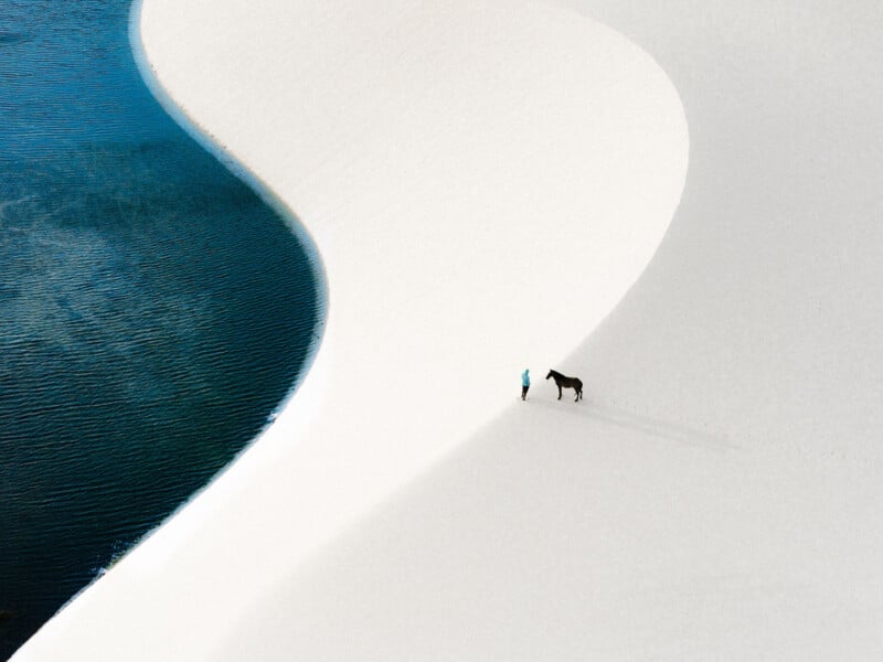 A person and a horse walk on white sand dunes beside a blue lagoon, creating a striking contrast between the smooth sand curves and the water.