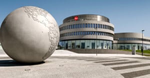 A large metallic globe sculpture stands on a plaza in front of a modern, circular building with a Leica logo and many windows under a clear blue sky.