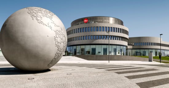 A large metallic globe sculpture stands on a plaza in front of a modern, circular building with a Leica logo and many windows under a clear blue sky.