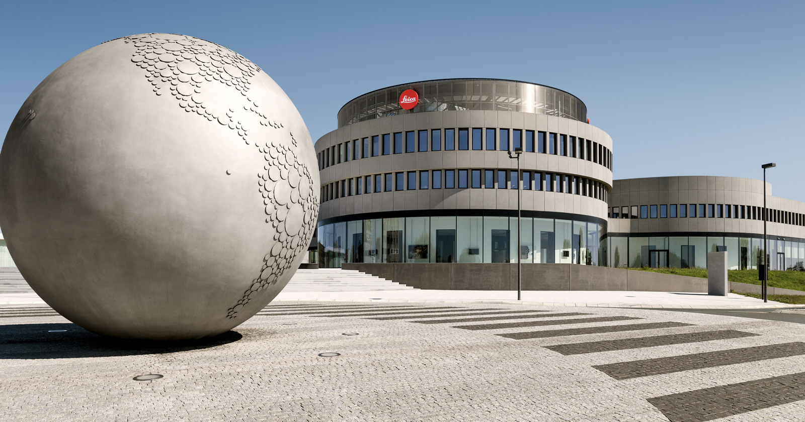 A large metallic globe sculpture stands on a plaza in front of a modern, circular building with a Leica logo and many windows under a clear blue sky.