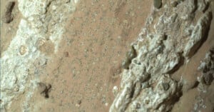 Close-up view of a rough, sandy surface with light brown and tan tones, scattered small stones, and irregular patches of white, textured material running diagonally across the image.