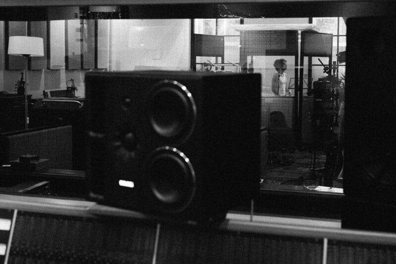 Foto en blanco y negro de Music Studio con un gran altavoz en primer plano, un hombre que está parado en una cabina insonorizada cantando o hablando en el micrófono en el fondo.