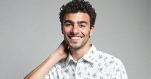 A young man with curly dark hair smiles at the camera, wearing a white shirt with small floral prints and holding one hand behind his neck against a plain, light gray background.