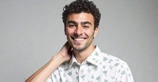 A young man with curly dark hair smiles at the camera, wearing a white shirt with small floral prints and holding one hand behind his neck against a plain, light gray background.