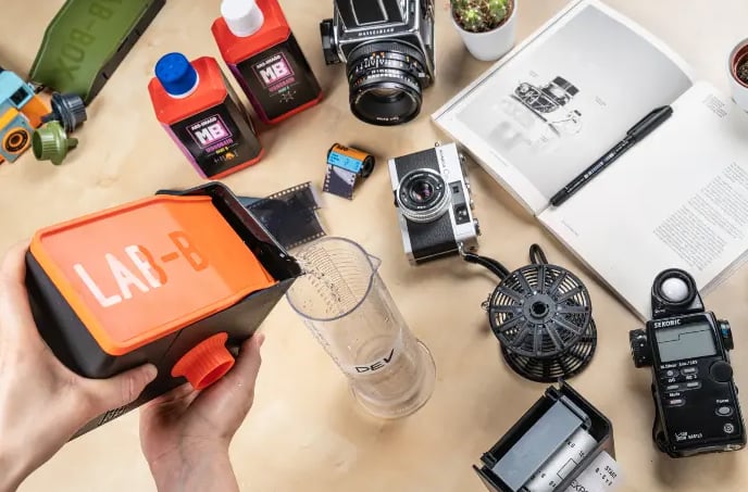 A person holds a LAB-BOX film developing tank on a table with cameras, film rolls, a developing reel, a beaker, light meter, pen, open book, and a small plant, showcasing film photography equipment.