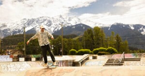 A person skateboards on the edge of a graffiti-covered skatepark ramp, with lush trees and snow-capped mountains in the background under a partly cloudy sky.