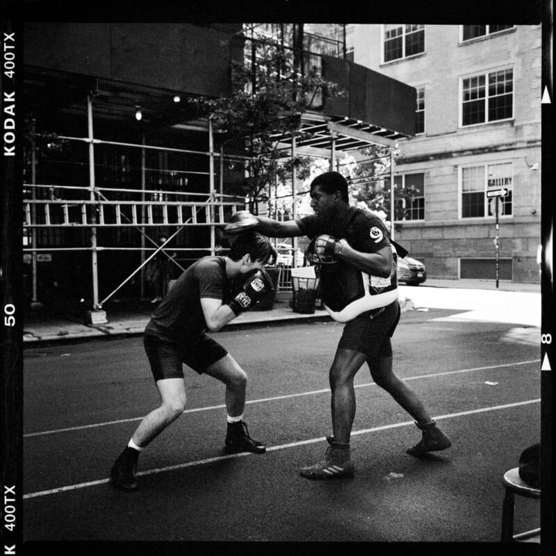 Two men box on a city street; one stands tall and extends a punch while the other crouches to dodge, both wearing boxing gloves and athletic gear. Scaffolding and buildings are visible in the background.