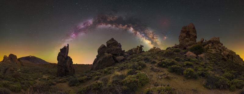 Rocky landscape with scattered green shrubs under a dark sky, featuring the Milky Way arched in bright colors above jagged rock formations at night.