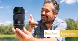 A smiling man outdoors holds up a large camera lens with his left hand and gestures toward it with his right. The logo "PetaPixel Showcase" appears in the lower right corner. Trees and a blue sky are in the background.