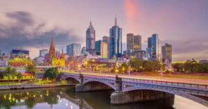 A cityscape of Melbourne at dusk, showing modern skyscrapers, historic buildings, streetlights, and a bridge over the Yarra River with reflections in the water.