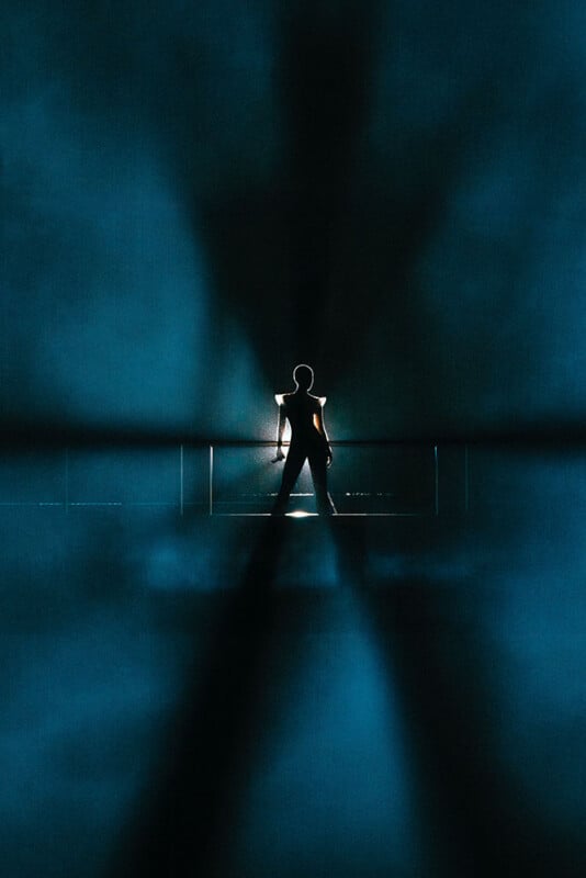 La silueta de una persona se encuentra con las piernas parcialmente abiertas, las manos en las caderas, retroiluminada por la luz brillante, creando sombras dramáticas y una atmósfera azul ahumada.