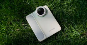 A white Littopo camera with a large circular lens lies on green grass, with sunlight highlighting the texture of both the device and the surrounding blades.