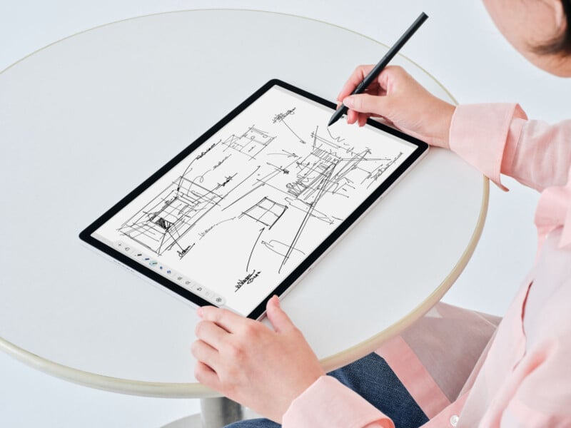 A person sketches architectural drawings on a digital tablet using a stylus while seated at a round white table. The tablet screen displays various building sketches and handwritten notes.