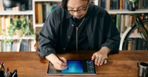 A person wearing glasses and dark clothing uses a stylus to draw on a tablet at a wooden desk, with bookshelves and a bicycle in the background.