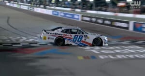 A white and blue racing car with the number 88 and WeatherTech branding crosses the finish line on a racetrack, with stands and lights visible in the background.