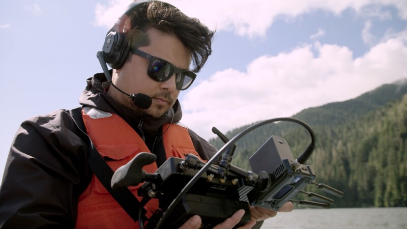 A man wearing sunglasses, a headset, and an orange life vest operates a remote control device outdoors, with a forested mountain and cloudy sky in the background.