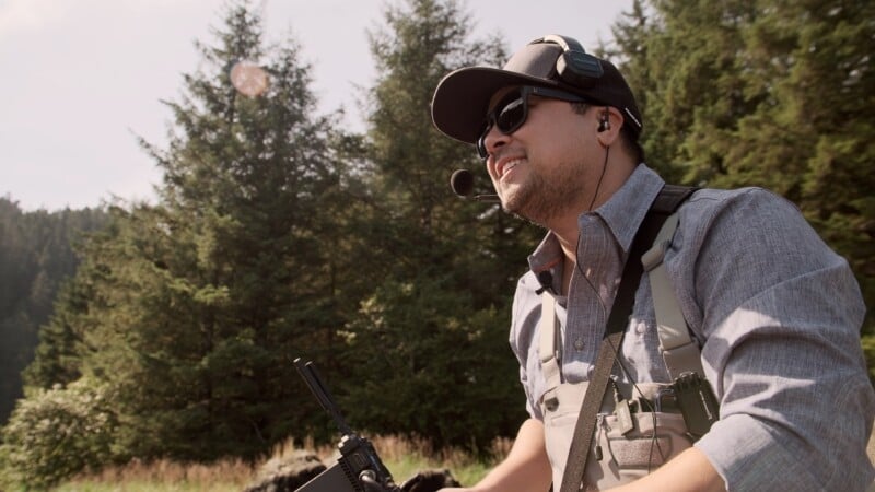 A man wearing sunglasses, a cap, and a headset speaks into a microphone outdoors, holding a walkie-talkie. He is surrounded by tall trees and sunlight.