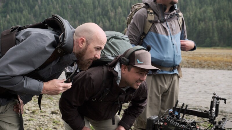Two men outdoors, wearing backpacks and outdoor clothing, closely examine a camera on a tripod near a rocky shore, with a forested area in the background. Another person stands nearby, partially out of frame.