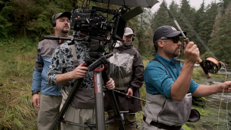Three men are outdoors near a forest; one operates a professional video camera on a tripod, while another fishes with a rod and reel. All wear outdoor gear, and the scene appears misty or rainy.