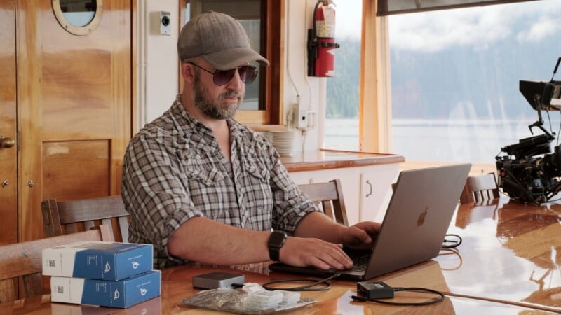 A bearded man wearing sunglasses, a gray cap, and a plaid shirt works on a laptop at a wooden table with electronic devices and boxes. He is inside a bright room with large windows and a fire extinguisher on the wall.