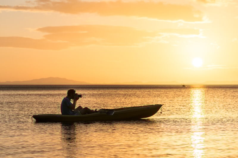 A man in a boat taking a picture of the sunset.