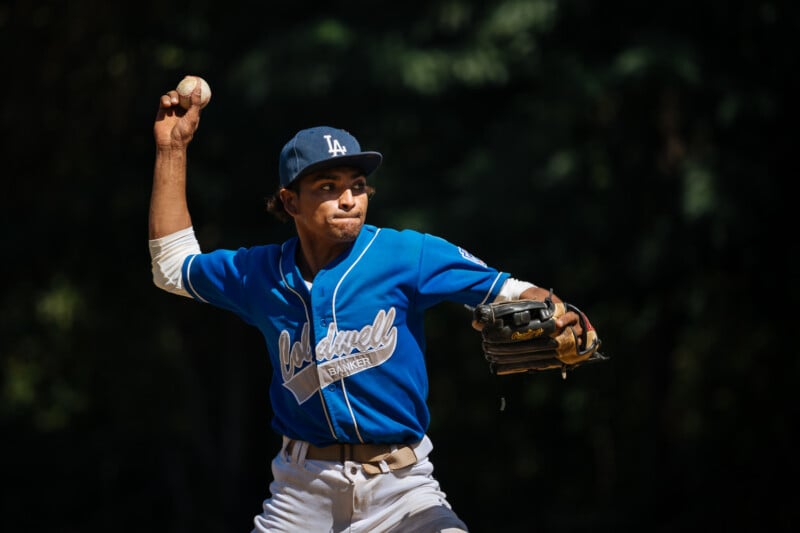 A baseball player in a blue uniform and cap prepares to throw a baseball, with his arm cocked back and intense focus on his face, set against a dark, blurred background.