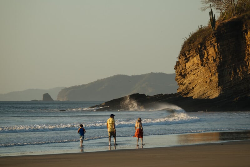 Three people—one adult and two children—stand on a sandy beach near the shoreline, looking toward the ocean. Rocky cliffs and waves are visible in the background under a clear sky.