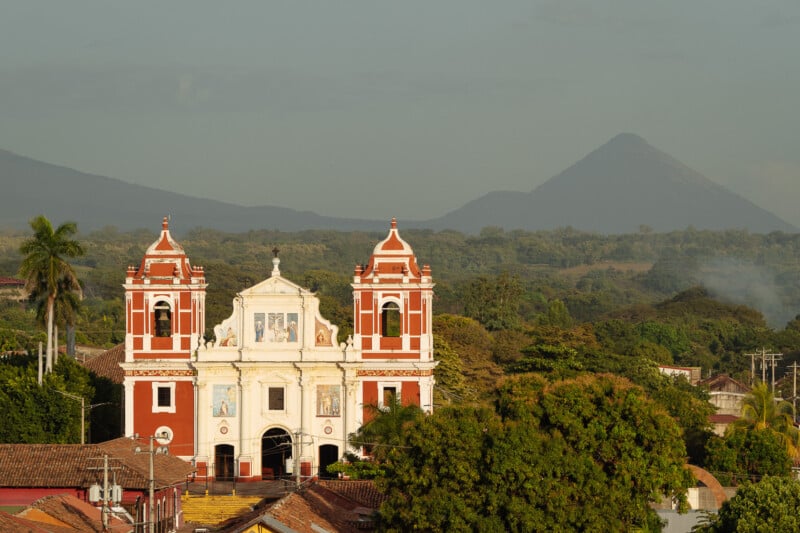A colonial-style church with red and white twin bell towers stands amid trees and rooftops, with lush green hills and a distant volcano in the background under a hazy sky.