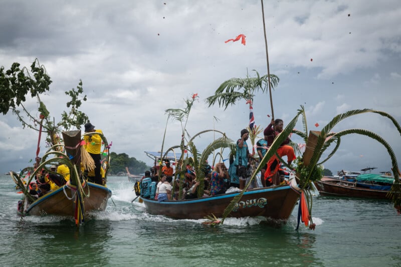 Two wooden boats decorated with palm fronds carry people dressed in colorful clothing, celebrating on the water under a cloudy sky. Some people throw items into the air, and other boats are visible in the background.